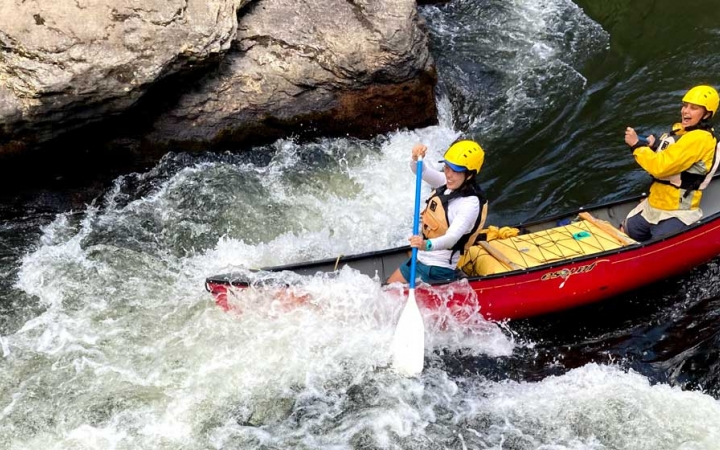 Two people wearing safety gear paddle a canoe through whitewater.